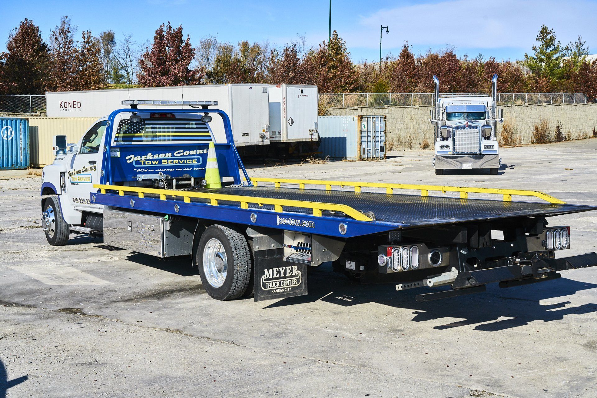 Blue-and-yellow flatbed tow truck parked in a gravel lot with shipping containers and trees in the background