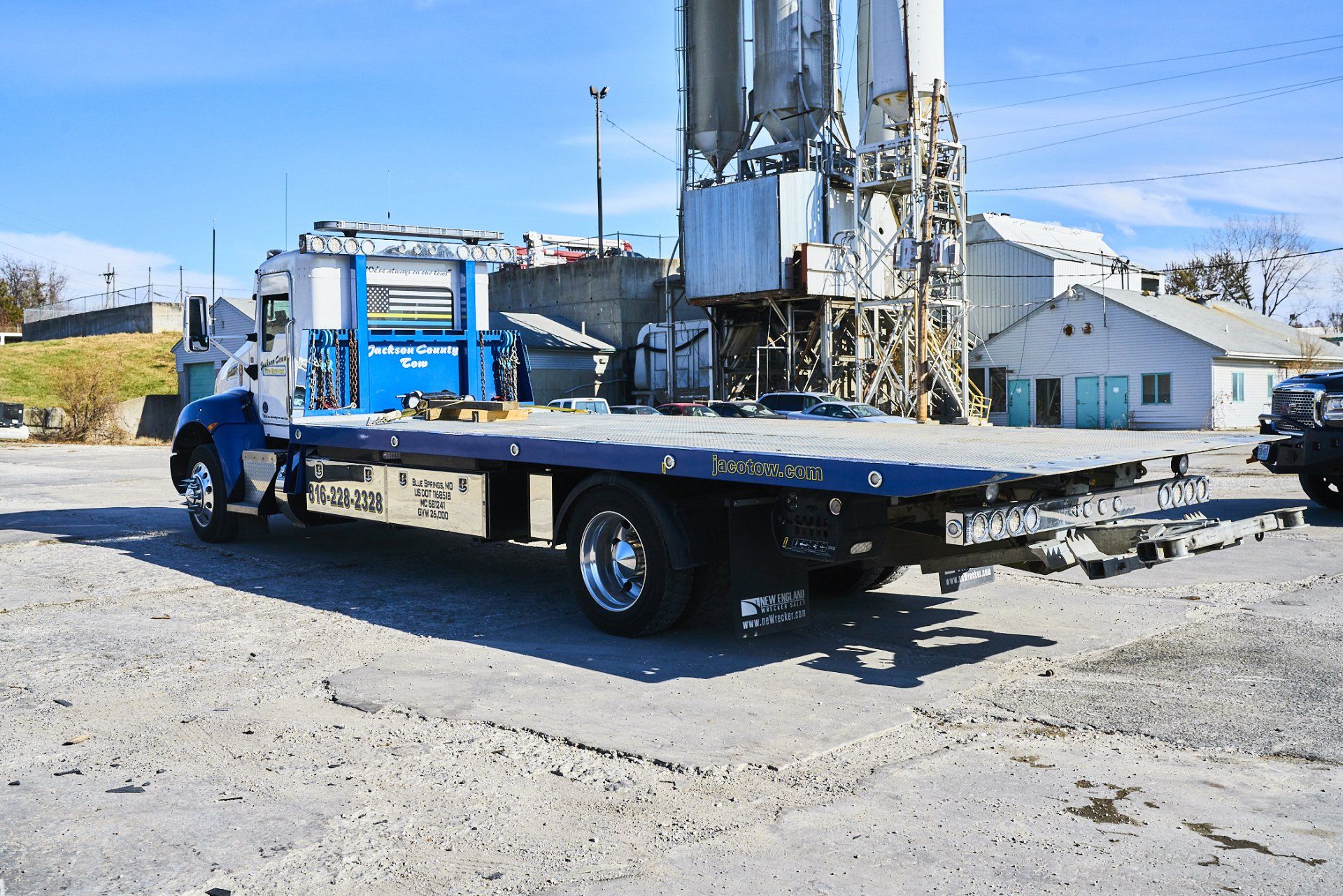 Blue tow truck carrying a drilling rig on a sunny roadside with buildings in the background