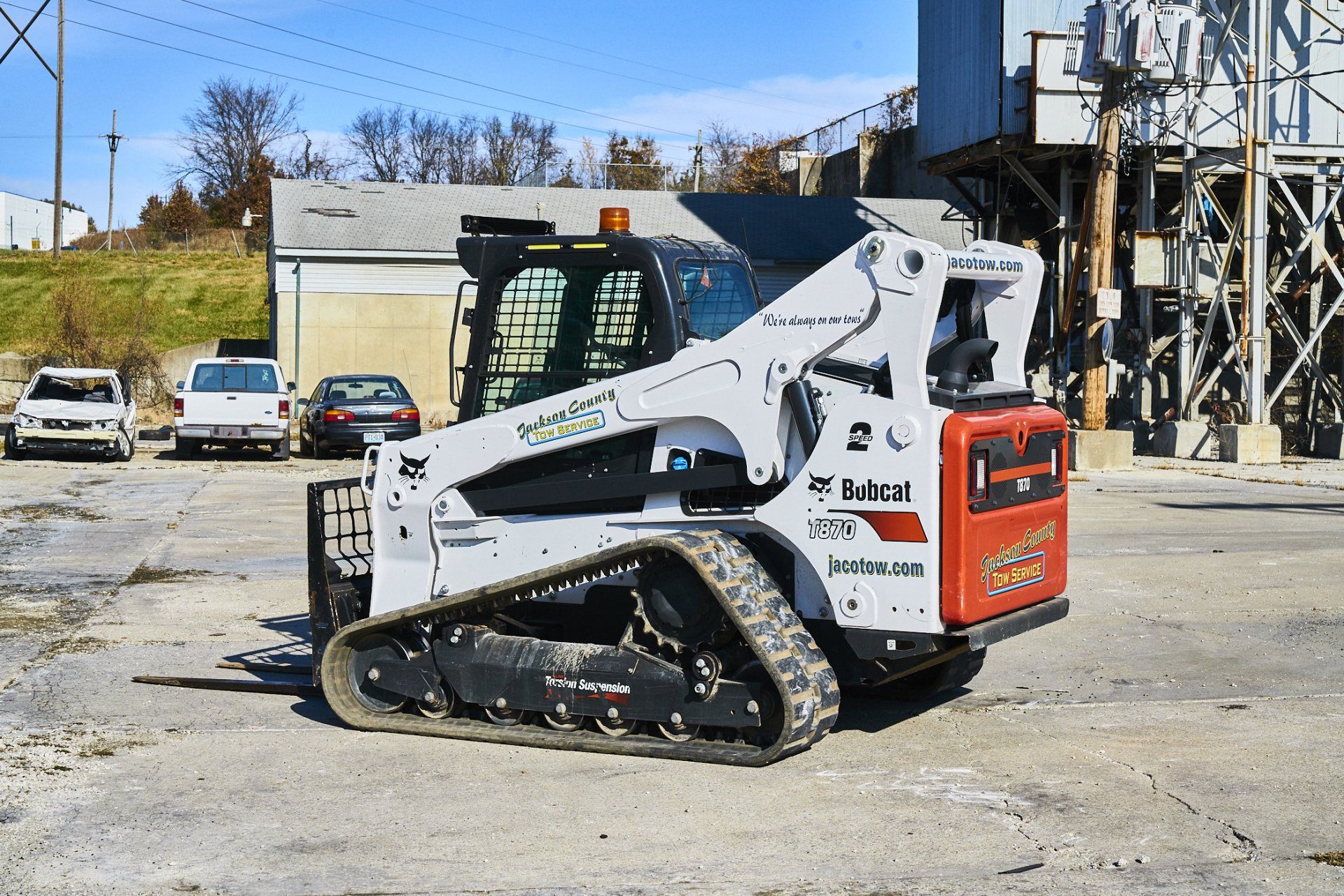 White Bobcat skid-steer loader parked in a gravel lot beside industrial equipment under a blue sky