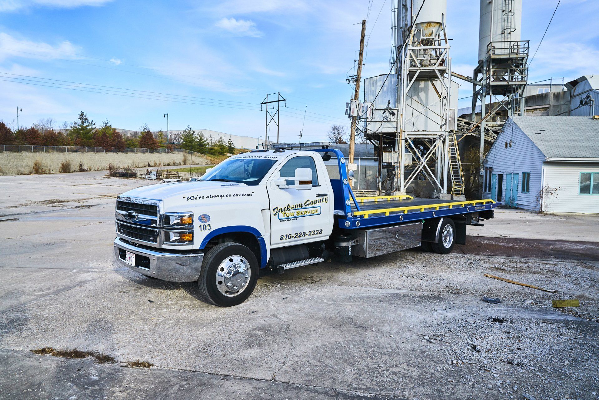 White flatbed tow truck parked on gravel beside an industrial building and silos