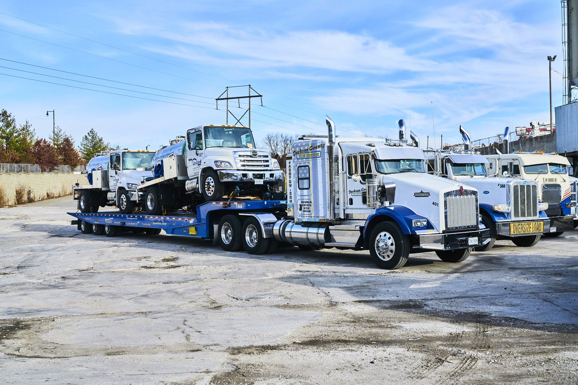 Flatbed tow truck hauling a white semi truck in a parking lot under a blue sky