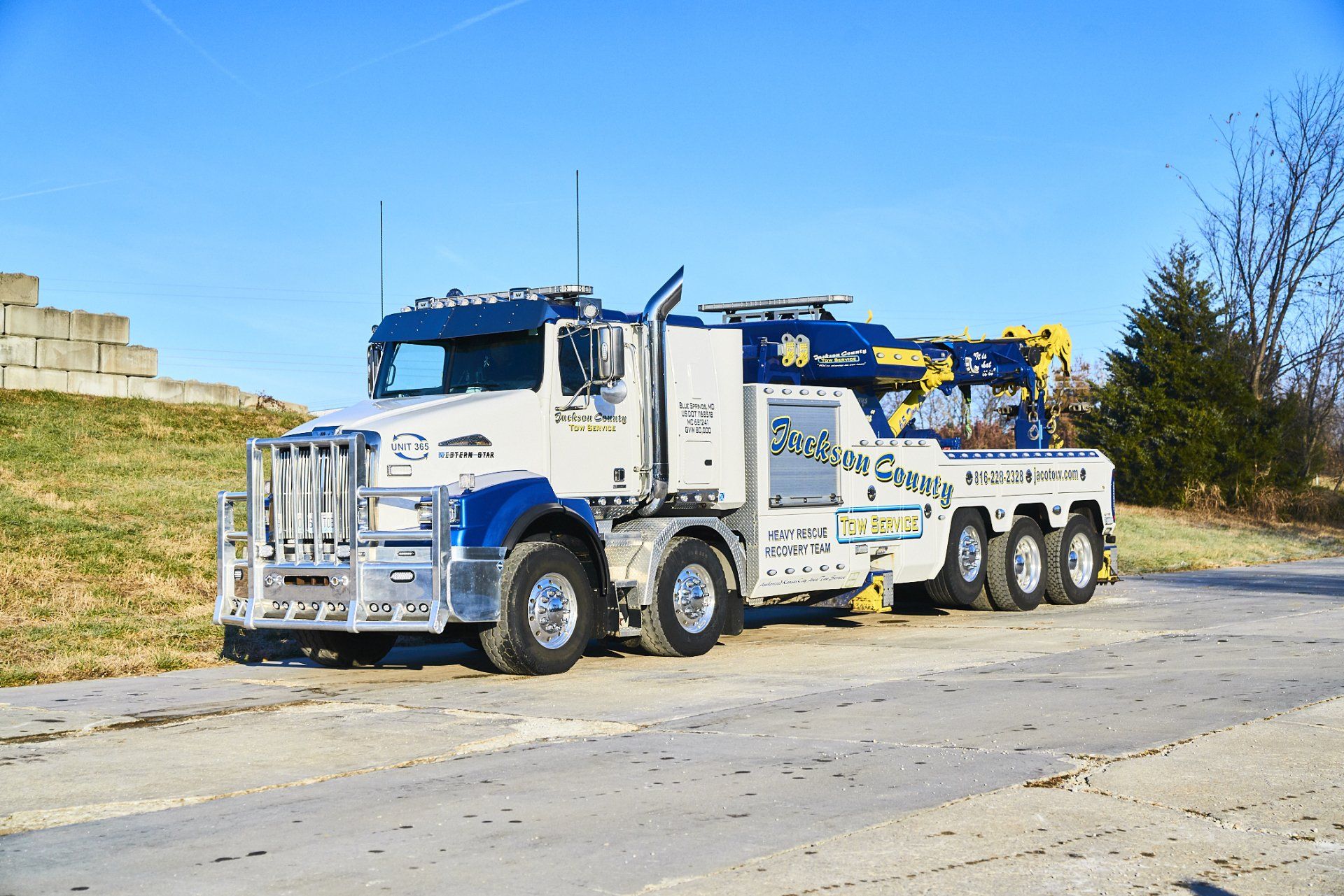 White tow truck with boom parked on a road beside a grassy embankment under a blue sky