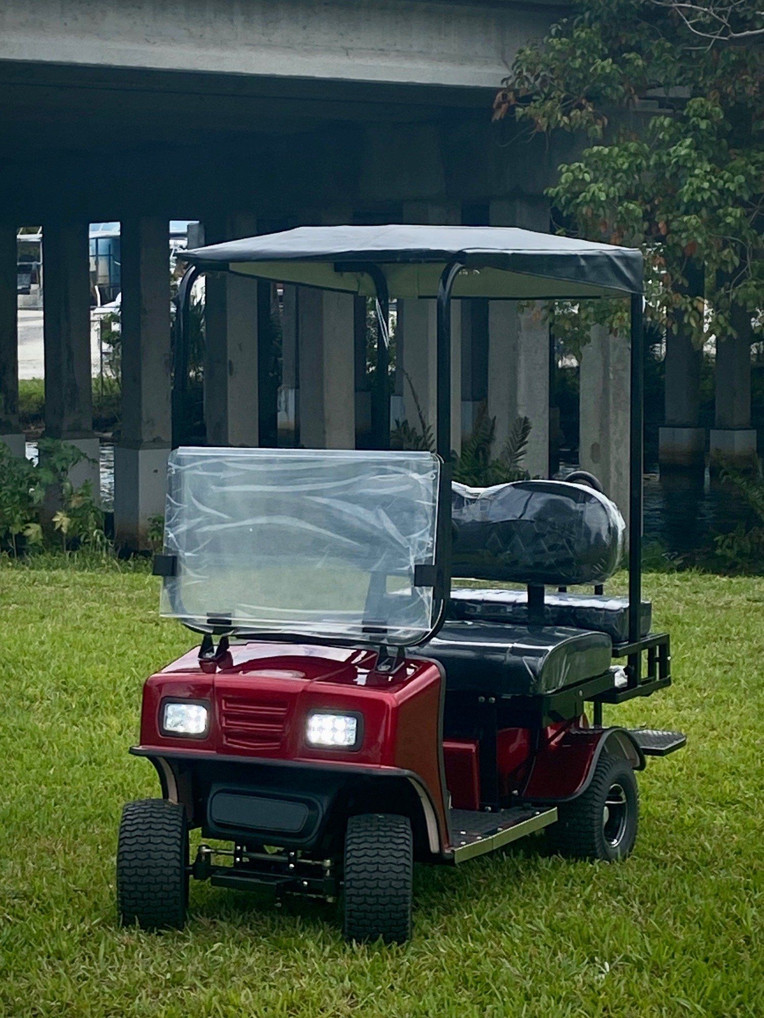 A red golf cart is parked in the grass under a bridge.