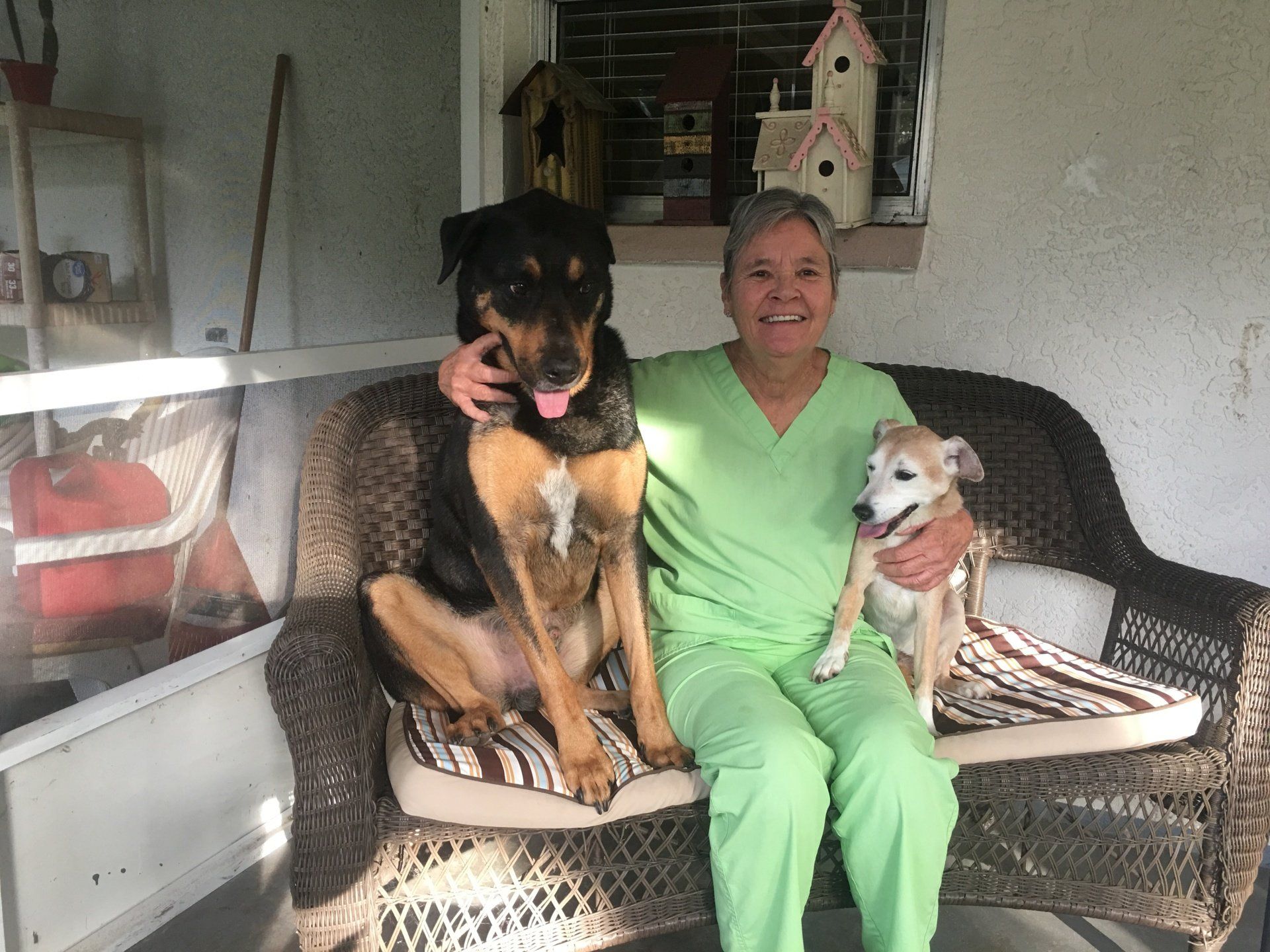 Woman in green scrubs with two dogs on a wicker bench outdoors.