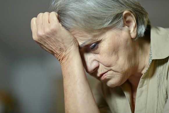 An elderly woman with gray hair, wearing a light brown shirt, looks down with a pensive expression, hand on forehead.