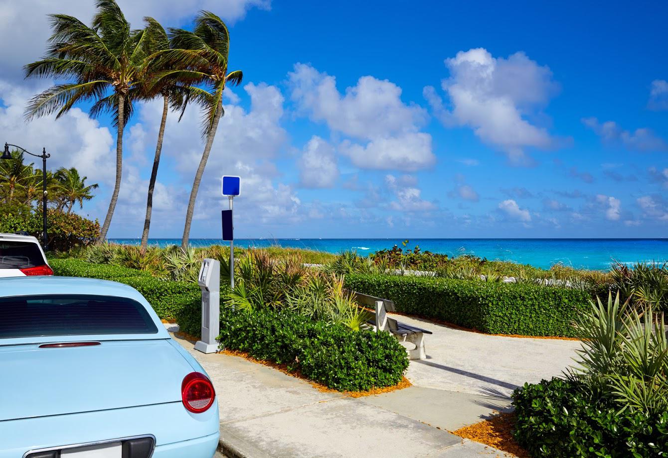 Seaside — Pensacola, FL — Matt McKeehan Bail Bonds Light blue car parked near a beach path, with palm trees, blue ocean, and sky with clouds in the background.