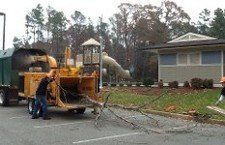 Tractor Mounted for Cutting a Tree — Williamsburg, VA — Top Notch Tree Service Workers feed tree branches into a yellow industrial wood chipper in a parking lot near a small building.