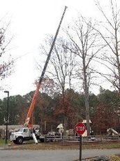 Crane — Williamsburg, VA — Top Notch Tree Service A crane truck with an extended boom parked at a playground, near a stop sign and leafless trees on an overcast day.