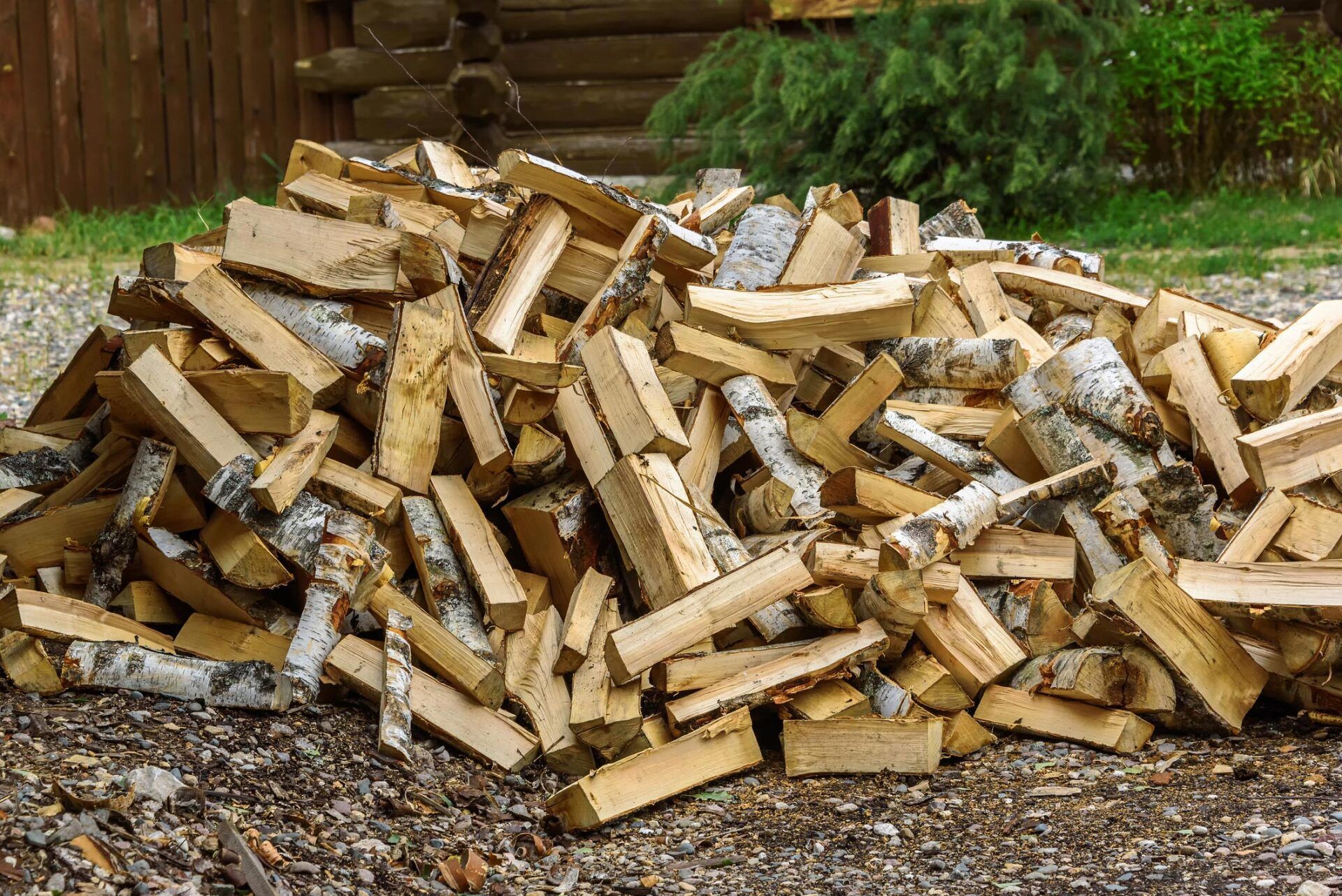 A large, messy pile of split birch firewood stacked on gravel ground in front of a log building.