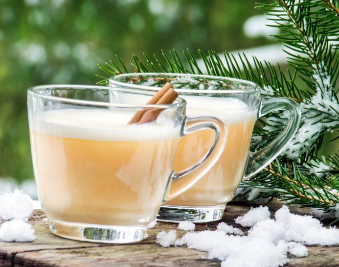 Zwei Glastassen mit gelbem Weihnachtspunsch und Zimtstange. Im Hintergrund verschneiter Tannenzweig