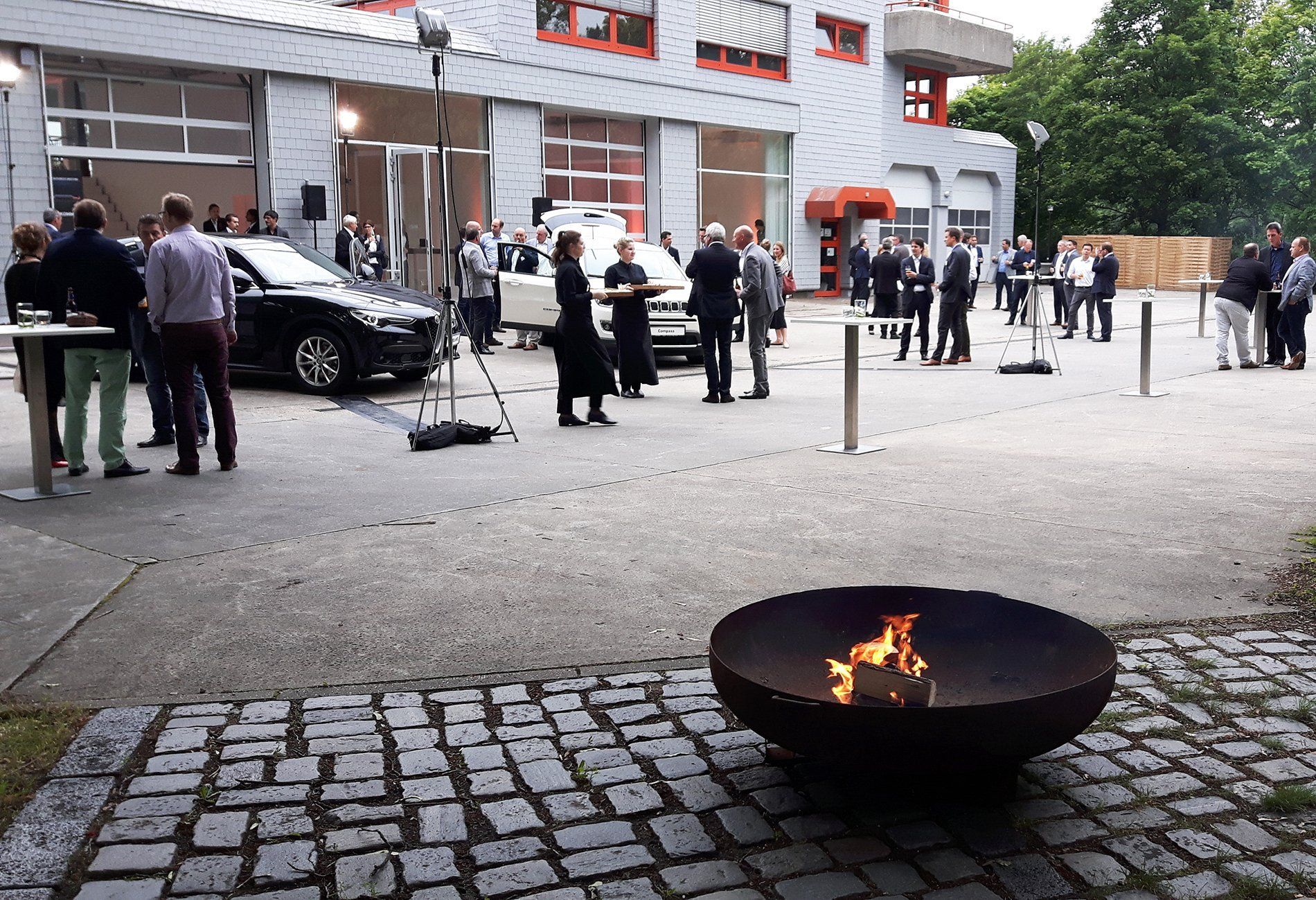 Feuerschale im Hof der Bridge Studios. Im HIntergrund Menschen und Range Rover Autos. Servicekräfte servieren Drinks