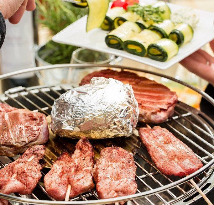 Rohe Steaks auf einem Grillrost. In der Mitte eine Backkartoffel in Alufolie