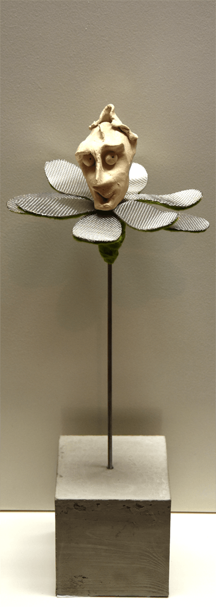 Plastik der Künstlerin Benita von Wendt, die eine Blume darstellt. In der Mitte der metallenen Blütenblätter sitzt ein flirtender menschlicher Kopf aus Ton. Der metallene Stil der Blume steckt auf einem quadratischen Betonsockel.