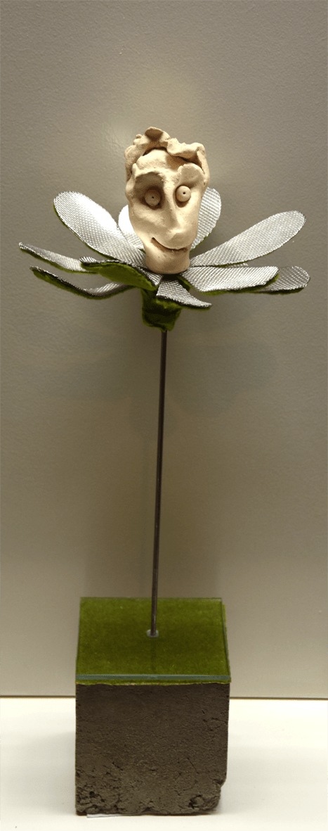 Plastik der Künstlerin Benita von Wendt, die eine Blume darstellt. In der Mitte der metallenen Blütenblätter sitzt ein lächelnder menschlicher Kopf aus Ton. Der metallene Stil der Blume steckt auf einem quadratischen Betonsockel.