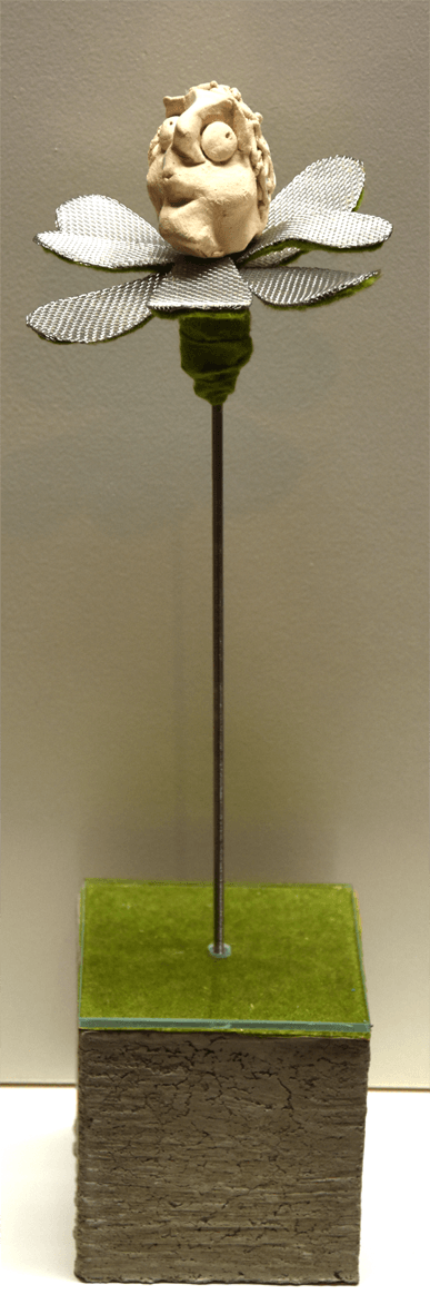 Plastik der Künstlerin Benita von Wendt, die eine Blume darstellt. In der Mitte der metallenen Blütenblätter sitzt ein pfeifender, betreten nach der Seite schauender menschlicher Kopf aus Ton. Der metallene Stil der Blume steckt auf einem quadratischen Betonsockel.