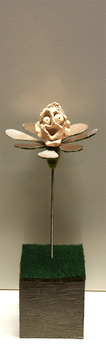 Plastik der Künstlerin Benita von Wendt, die eine Blume darstellt. In der Mitte der metallenen Blütenblätter sitzt ein lachender menschlicher Kopf aus Ton. Der metallene Stil der Blume steckt auf einem quadratischen Betonsockel.