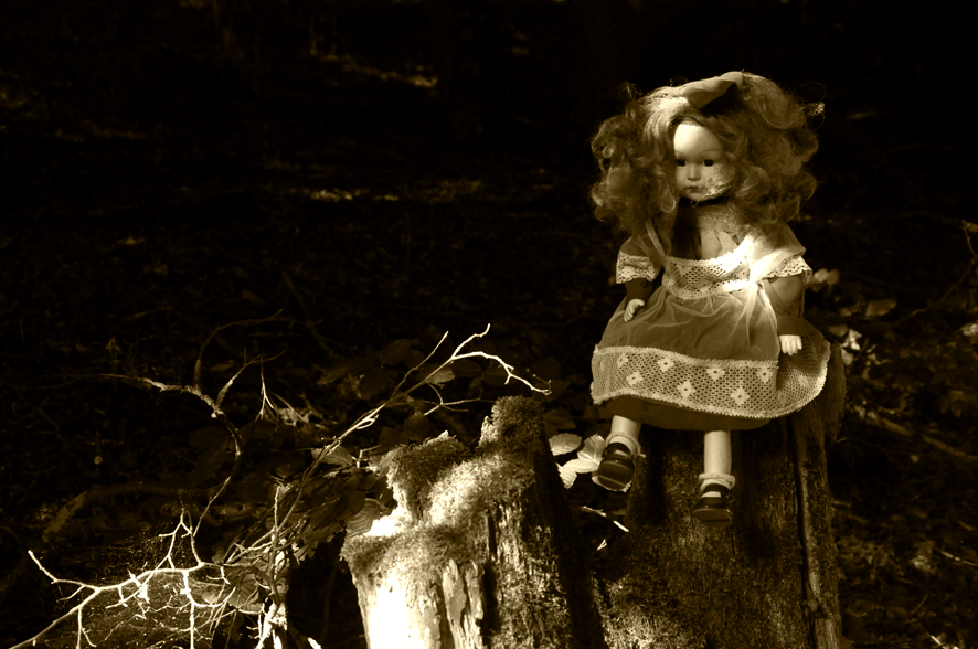 Fotografie der Künstlerin Benita von Wendt. Titel: Einsamkeit. Die Fotografie zeigt eine weibliche altmodische Puppe die auf einem Baumstumpf sitzt. Die Fotografie ist in Sepiatönen gehalten. Ein Spiel aus Licht und Schatten setzt die Puppe in Szene, die Umgebung löst sich in der Dunkelheit auf.