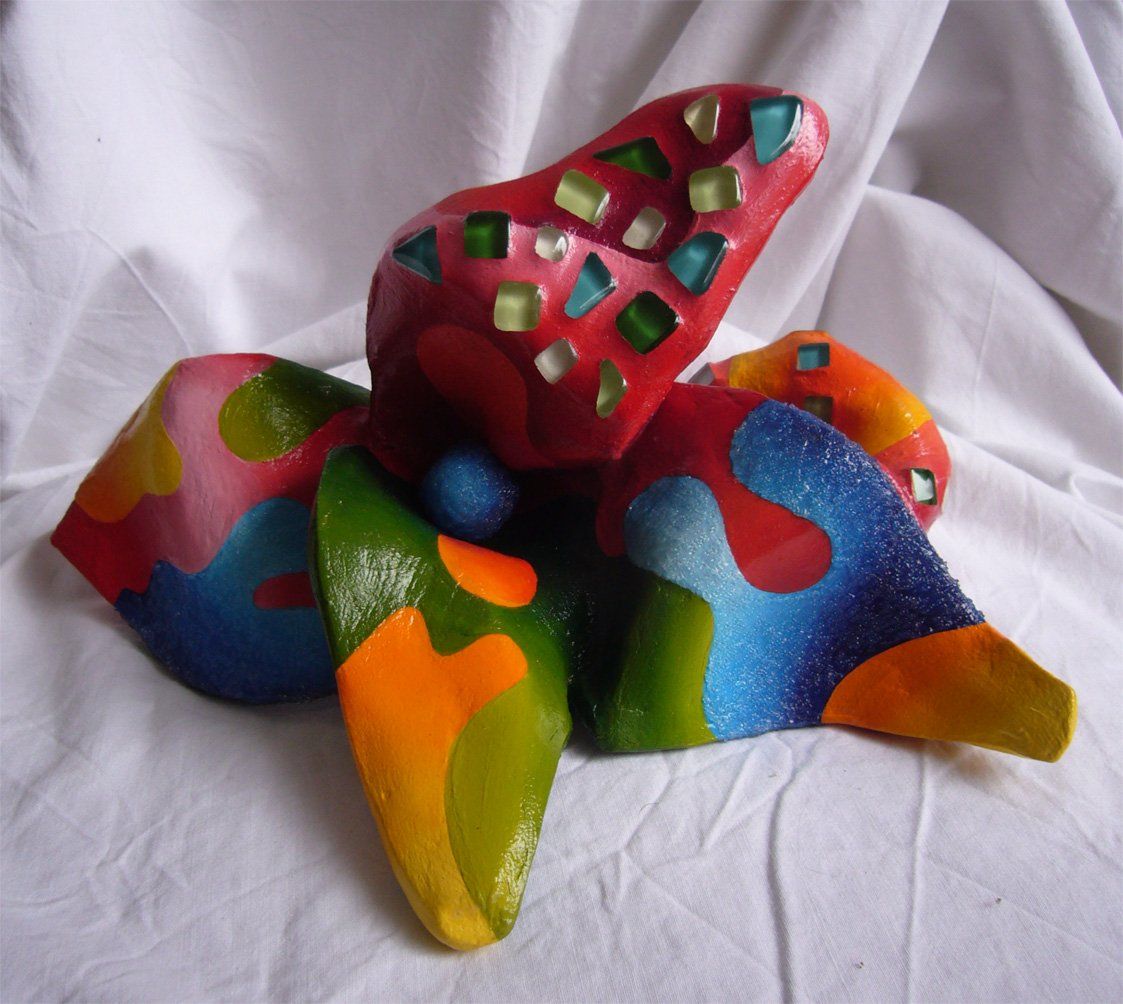 Plastik aus Pappmasché der Künstlerin Benita von Wendt. Titel: Fleur d'espoir 1. Bunte Skulptur die eine kleine vierblätterige Blume, die auf dem Boden liegt darstellt. Die Blume ist mit bunten, abstrakten Ornamenten bemalt und mit Mosaiksteinen besetzt.