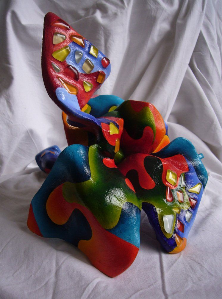 Plastik aus Pappmasché der Künstlerin Benita von Wendt. Titel: Fleur d'espoir 3. Bunte Skulptur die eine kleine sechsblätterige Blume, die auf dem Boden liegt darstellt. Die Blume ist mit bunten, abstrakten Ornamenten bemalt und mit Mosaiksteinen besetzt. Seitenansicht rechts.