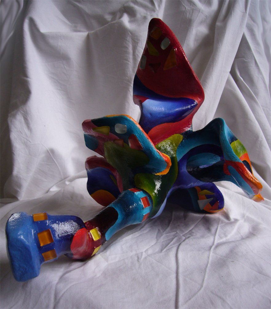 Plastik aus Pappmasché der Künstlerin Benita von Wendt. Titel: Fleur d'espoir 3. Bunte Skulptur die eine kleine sechsblätterige Blume, die auf dem Boden liegt darstellt. Die Blume ist mit bunten, abstrakten Ornamenten bemalt und mit Mosaiksteinen besetzt. Rückansicht.