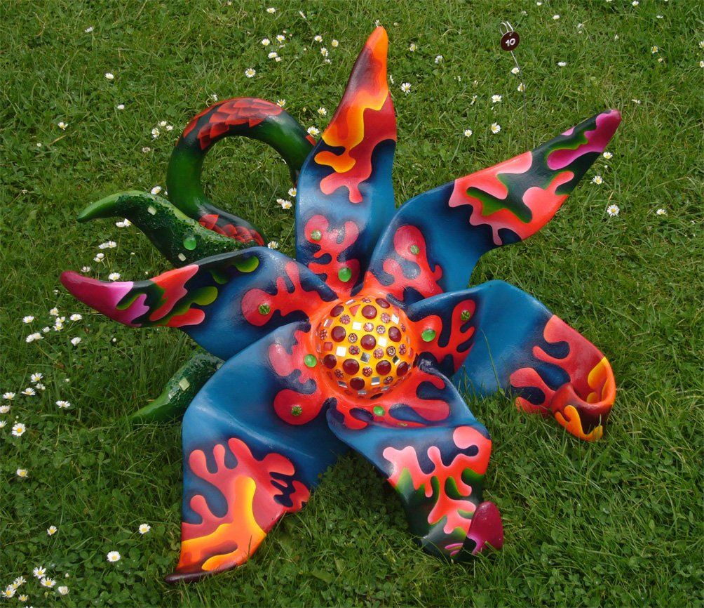 Plastik aus Pappmasché der Künstlerin Benita von Wendt. Titel: Blume im Gras. Bunte Skulptur die eine große Blume, die auf dem Boden liegt darstellt. Die Blume besteht aus einem Blütenstempel mit Mosaiksteinen besetzt, türkisen Blütenblättern mit abstrakten bunten Ornamenten, sowie einem grünen Stiel der mit Laub ähnlichen Formen bemalt ist. Frontansicht.