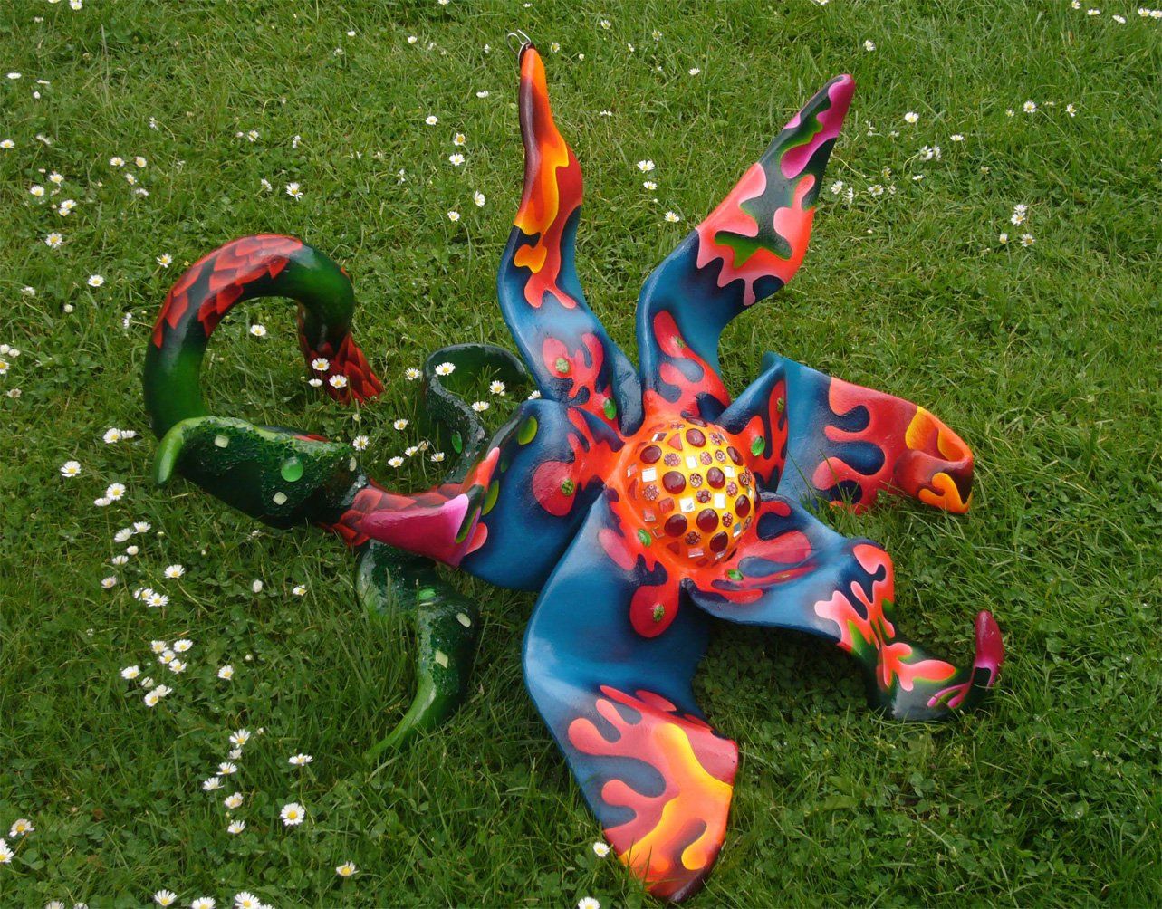 Plastik aus Pappmasché der Künstlerin Benita von Wendt. Titel: Blume im Gras. Bunte Skulptur die eine große Blume, die auf dem Boden liegt darstellt. Die Blume besteht aus einem Blütenstempel mit Mosaiksteinen besetzt, türkisen Blütenblättern mit abstrakten bunten Ornamenten, sowie einem grünen Stiel der mit Laub ähnlichen Formen bemalt ist. Seitenansicht.