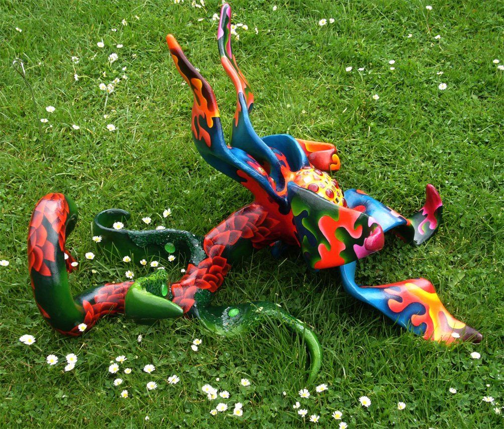 Plastik aus Pappmasché der Künstlerin Benita von Wendt. Titel: Blume im Gras. Bunte Skulptur die eine große Blume, die auf dem Boden liegt darstellt. Die Blume besteht aus einem Blütenstempel mit Mosaiksteinen besetzt, türkisen Blütenblättern mit abstrakten bunten Ornamenten, sowie einem grünen Stiel der mit Laub ähnlichen Formen bemalt ist. Seitenansicht halblinks..