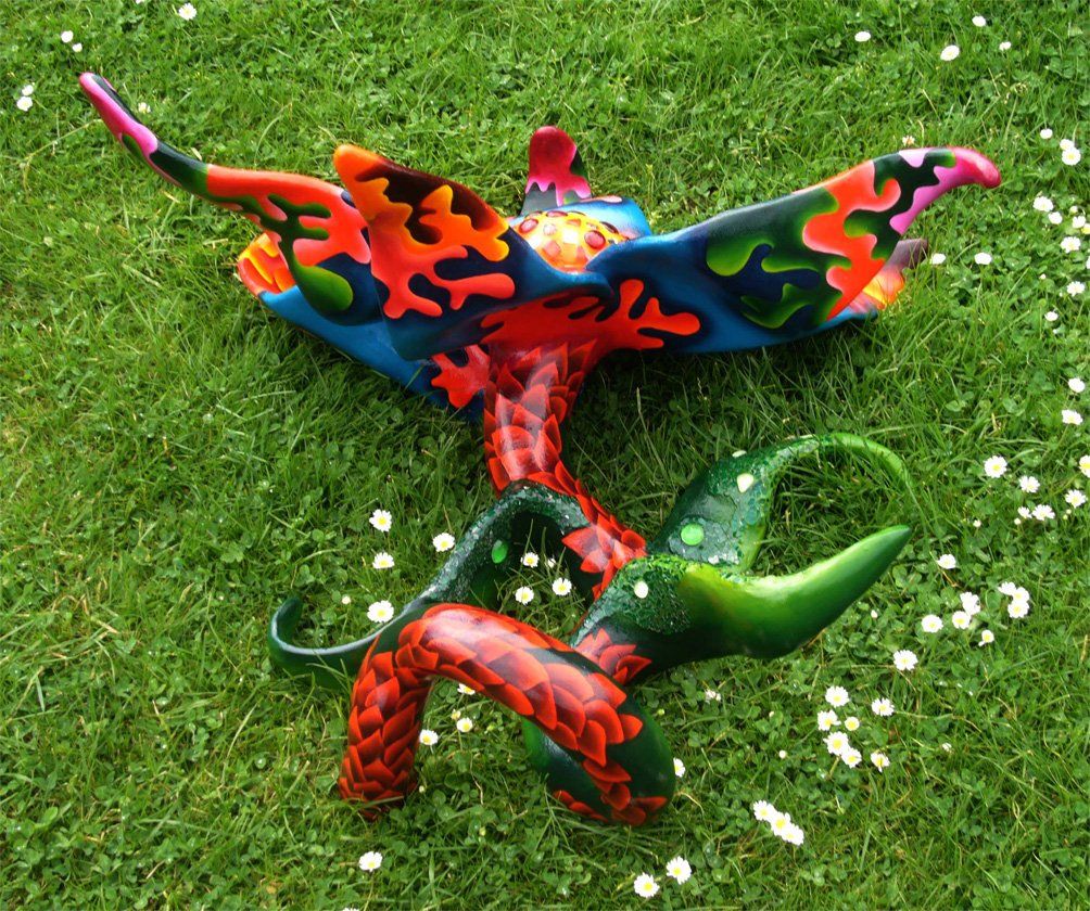 Plastik aus Pappmasché der Künstlerin Benita von Wendt. Titel: Blume im Gras. Bunte Skulptur die eine große Blume, die auf dem Boden liegt darstellt. Die Blume besteht aus einem Blütenstempel mit Mosaiksteinen besetzt, türkisen Blütenblättern mit abstrakten bunten Ornamenten, sowie einem grünen Stiel der mit Laub ähnlichen Formen bemalt ist. Rückansicht.