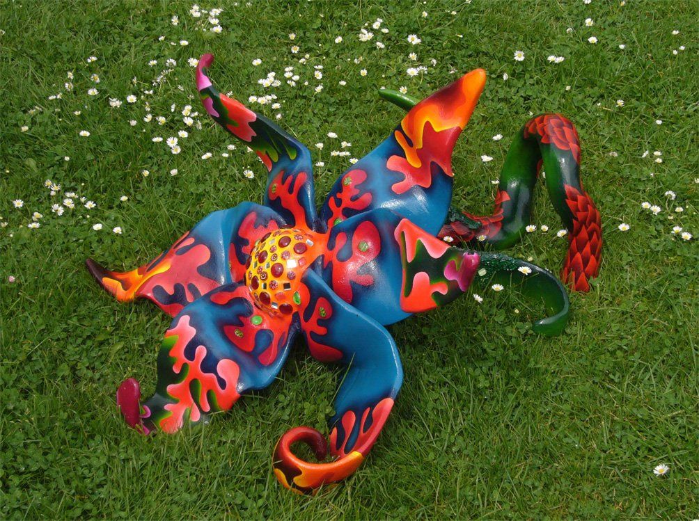 Plastik aus Pappmasché der Künstlerin Benita von Wendt. Titel: Blume im Gras. Bunte Skulptur die eine große Blume, die auf dem Boden liegt darstellt. Die Blume besteht aus einem Blütenstempel mit Mosaiksteinen besetzt, türkisen Blütenblättern mit abstrakten bunten Ornamenten, sowie einem grünen Stiel der mit Laub ähnlichen Formen bemalt ist.