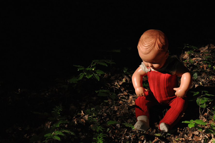 Fotografie der Künstlerin Benita von Wendt. Titel: Gegenstand, männlich N° 209. Die Fotografie zeigt eine männliche altmodische Puppe die auf dem Waldboden sitzt, ein Spiel aus Licht und Schatten setzt die Puppe in Szene, die Umgebung löst sich in der Dunkelheit auf.
