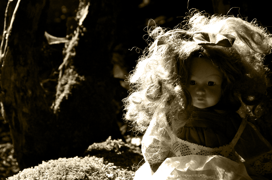 Fotografie der Künstlerin Benita von Wendt. Titel: Zweifel. Die Fotografie zeigt eine weibliche altmodische Puppe die auf einem moosbewachsenen Stein sitzt. Die Fotografie ist in Sepiatönen gehalten. Ein Spiel aus Licht und Schatten setzt die Puppe in Szene, die Umgebung löst sich in der Dunkelheit auf.