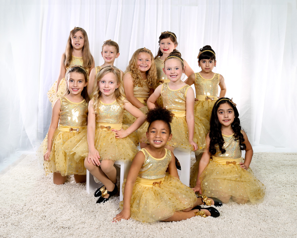 A group of young girls in gold dresses, smiling, posed on a rug in front of white curtains.