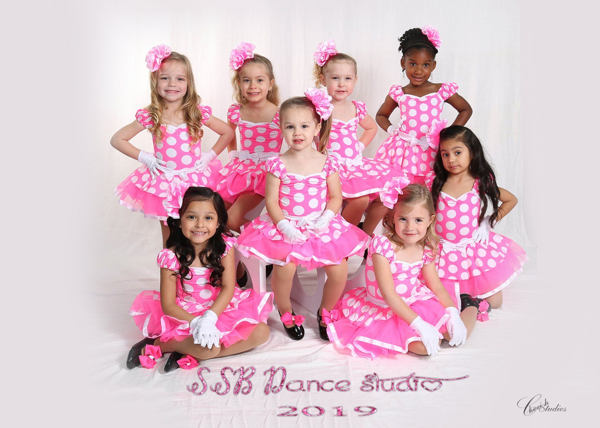 Group of young girls in pink polka-dot dance costumes, smiling, posing for a studio photo.