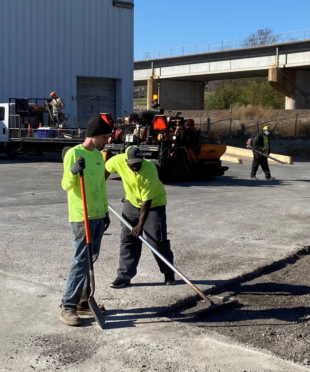 Workers in neon safety shirts paving gravel near construction equipment beside an overpass.