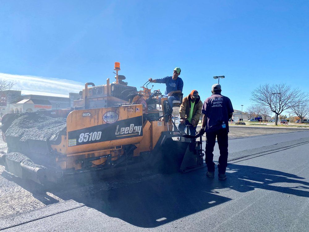 Road crew repairing asphalt around an orange Leeboy paving machine on a sunny street