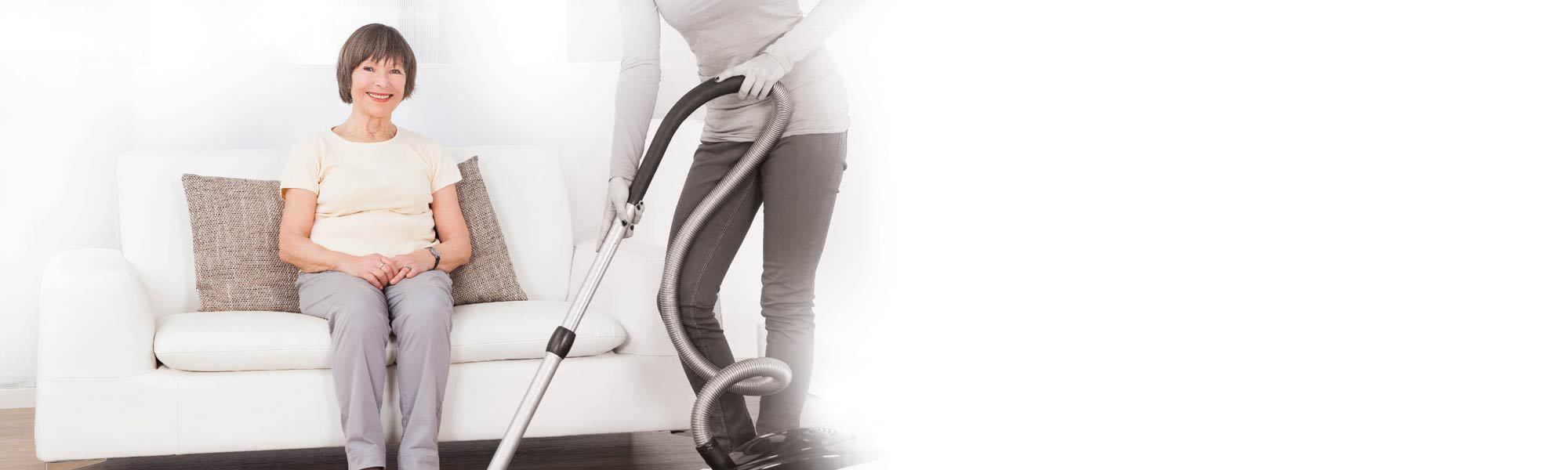 caretaker cleaning floor with vacuum cleaner while senior woman sitting on sofa at nursing home