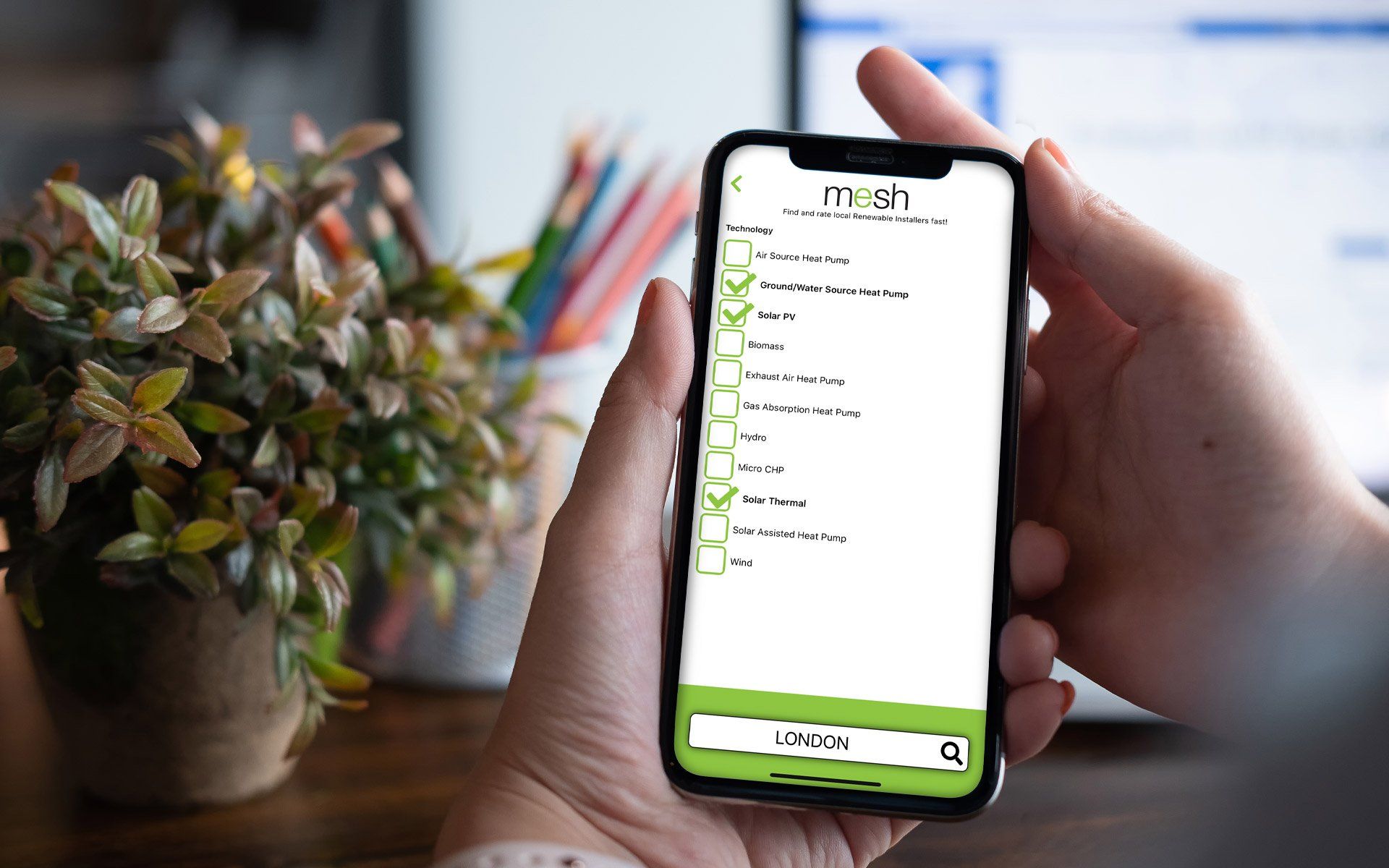 Hands holding a smartphone showing a green to-do list app beside a potted plant and colored pencils