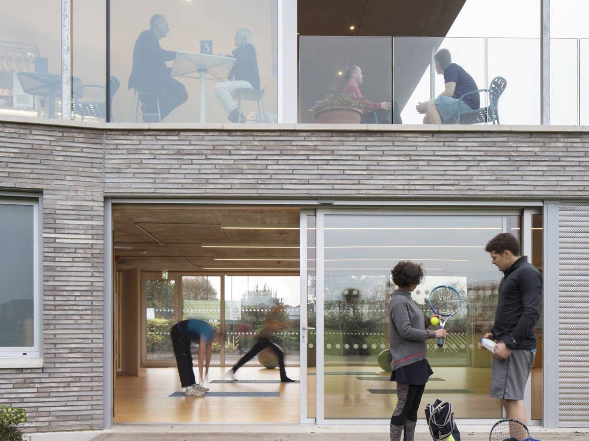 People talking outside a modern house while others exercise inside through large windows