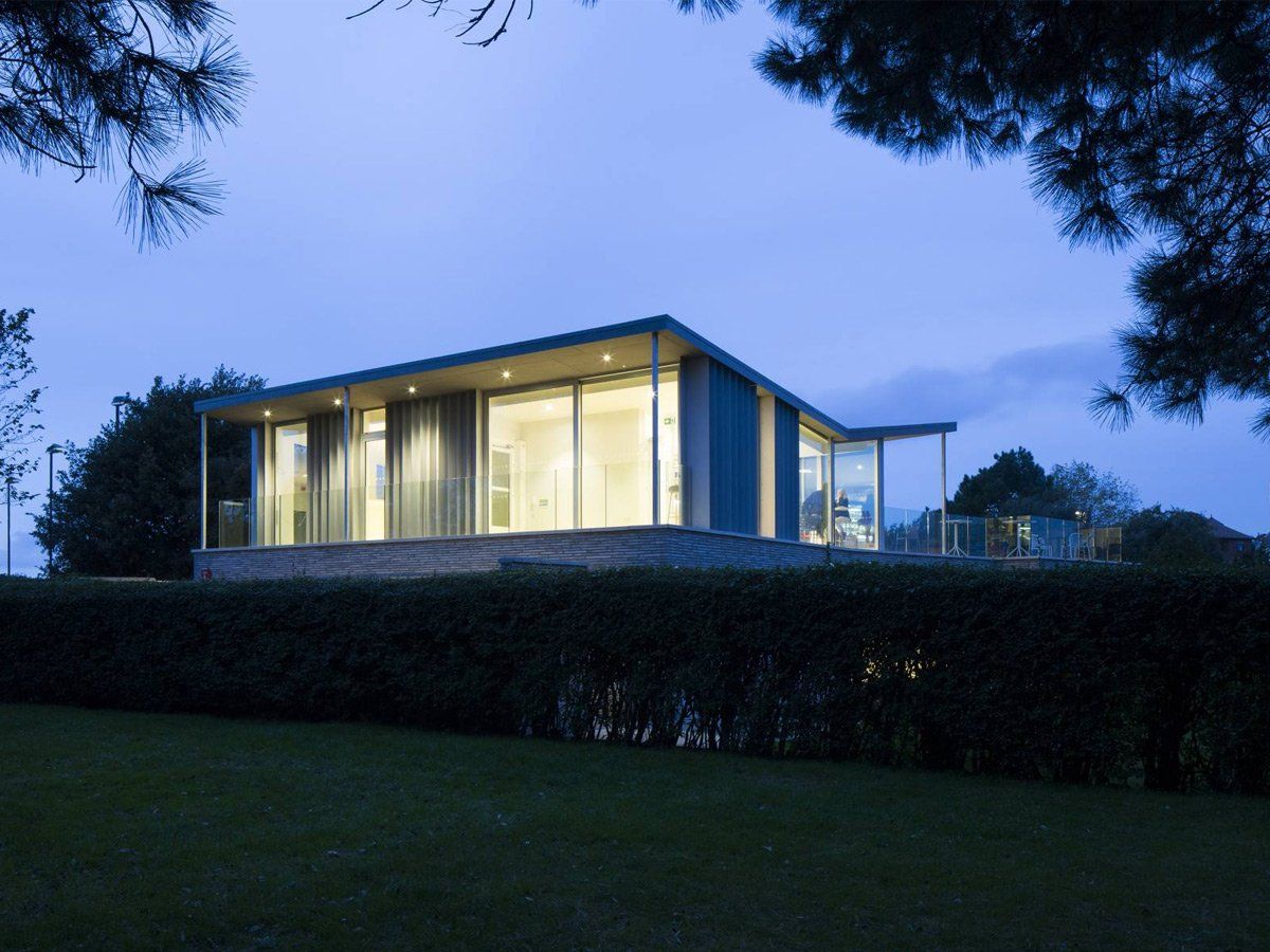 Modern glass house at dusk, warmly lit behind a hedge with dark trees framing the scene
