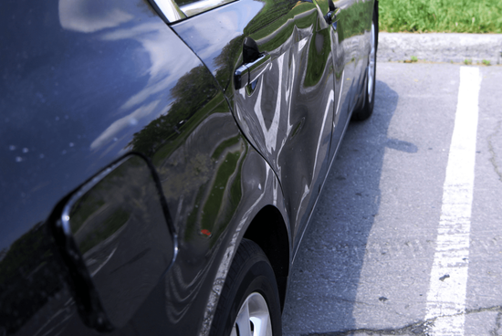 Damaged black car side panel in a parking lot, with a large dent near the rear wheel.