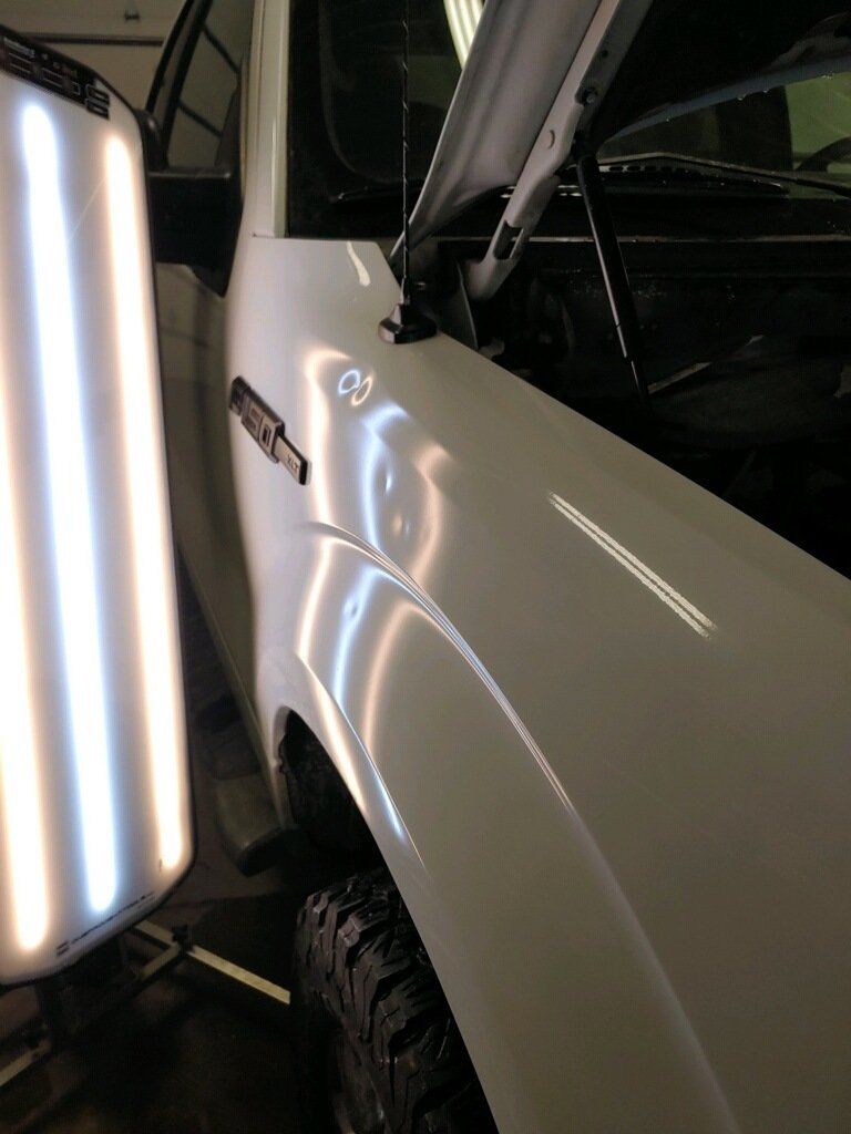 Close-up of a white truck’s side mirror and front fender under bright garage lights