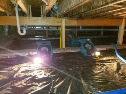 Two workers under a house crawlspace repairing pipes with flashlights and tools on a plastic-covered floor