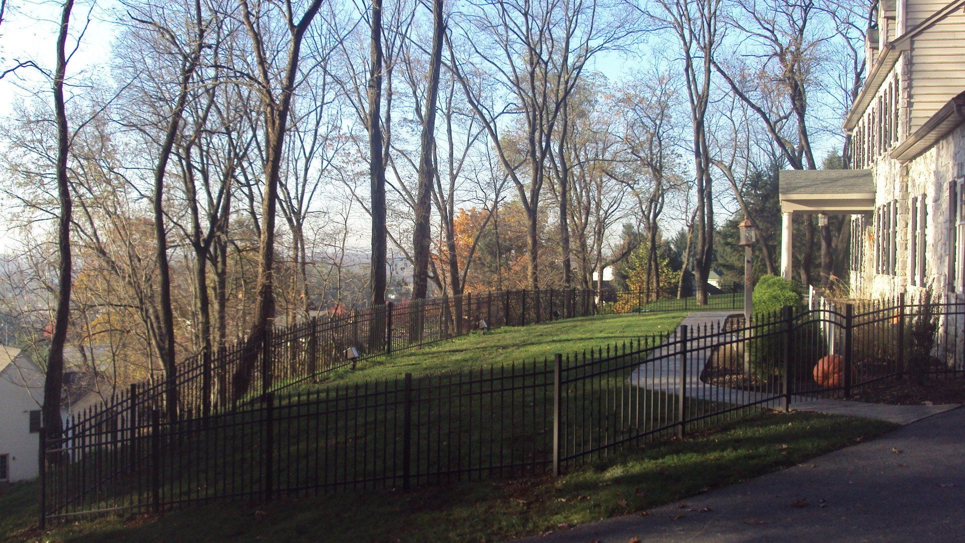 Black metal fence bordering a grassy hill with trees and a house on a sunny day.