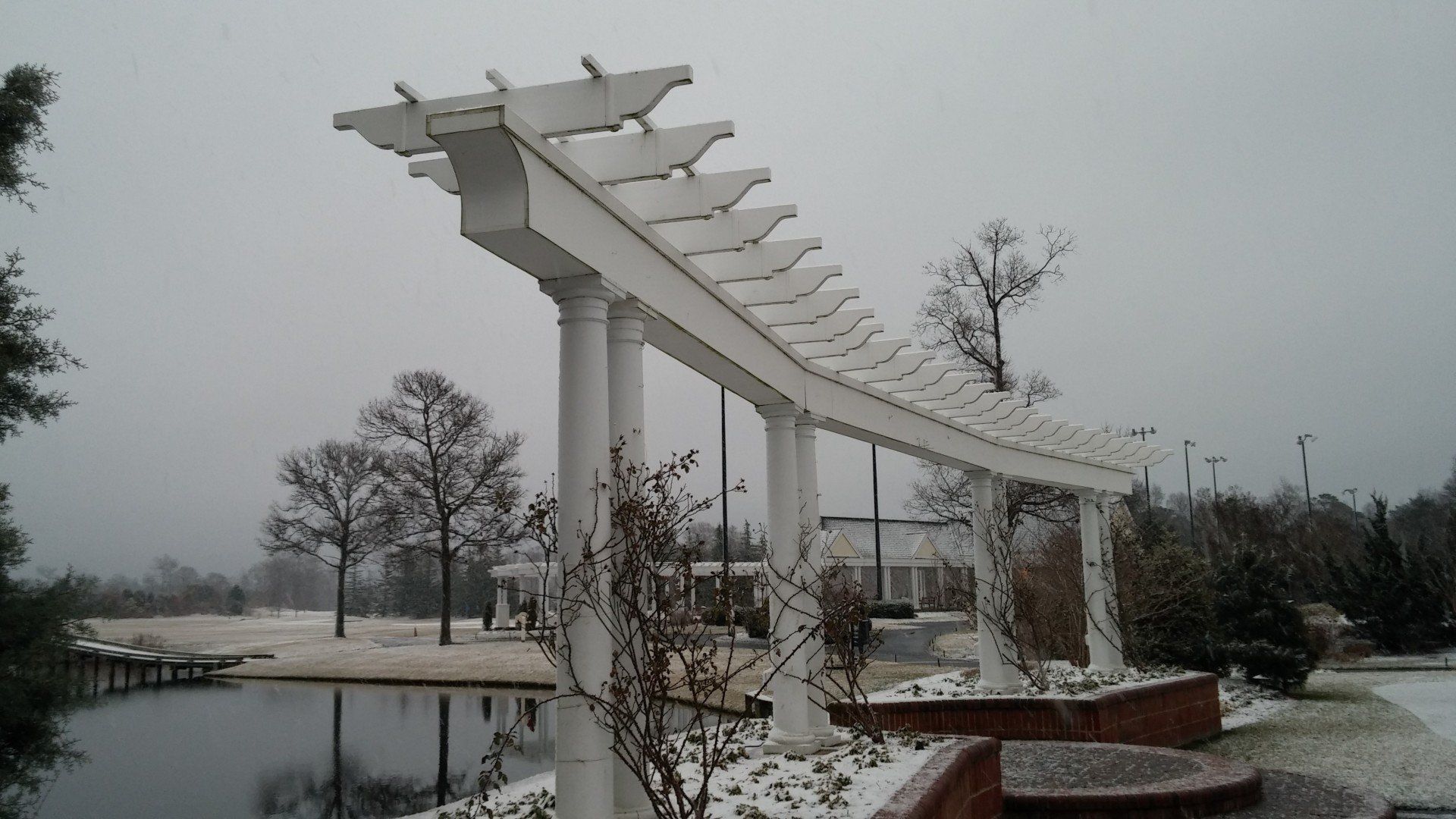 White pergola in a snowy landscape, over a pond. Bare trees, overcast sky, snow-covered ground.