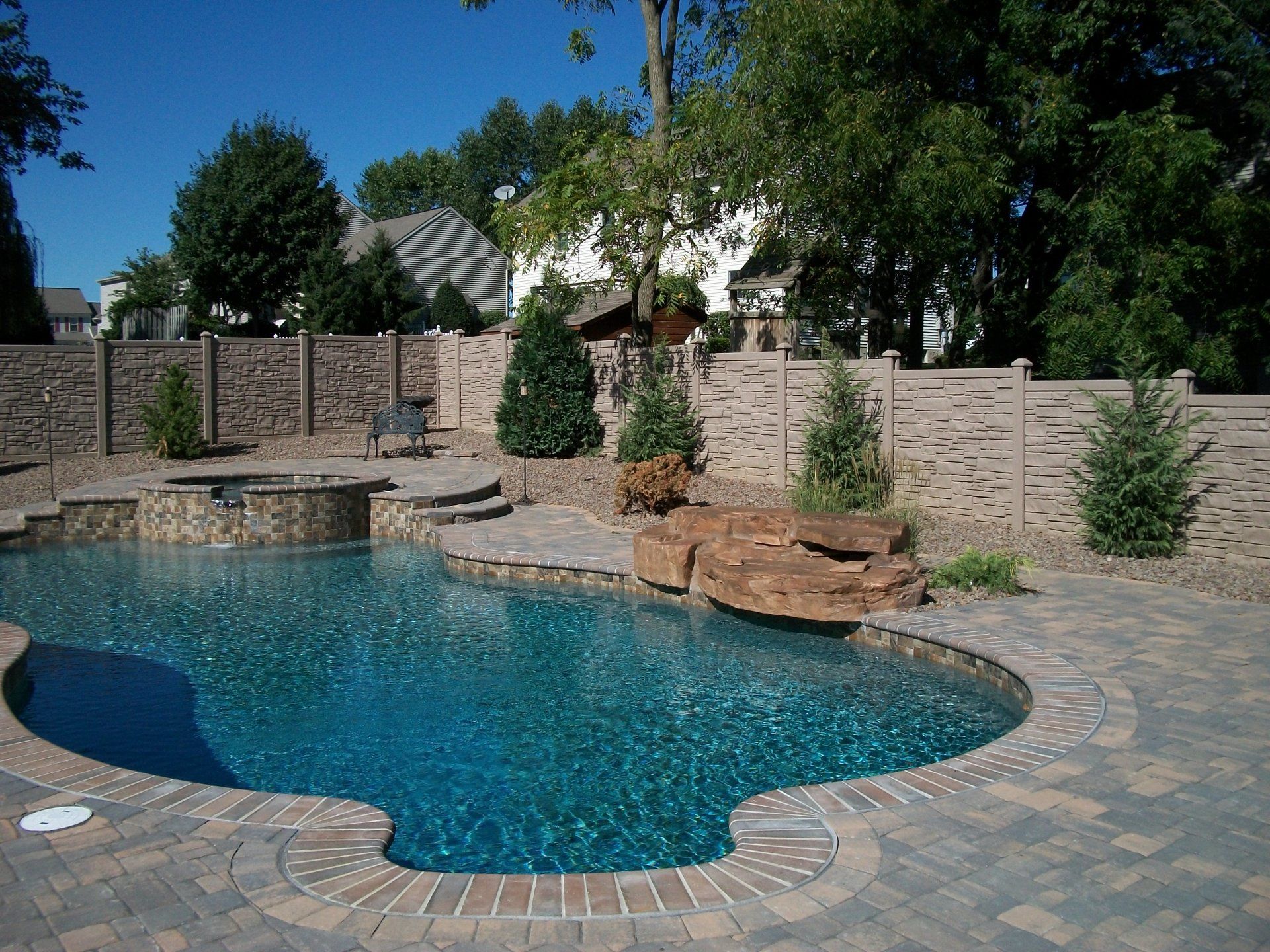 Swimming pool with stone surround, a hot tub, and a brick fence in a backyard setting in Lititz, PA.