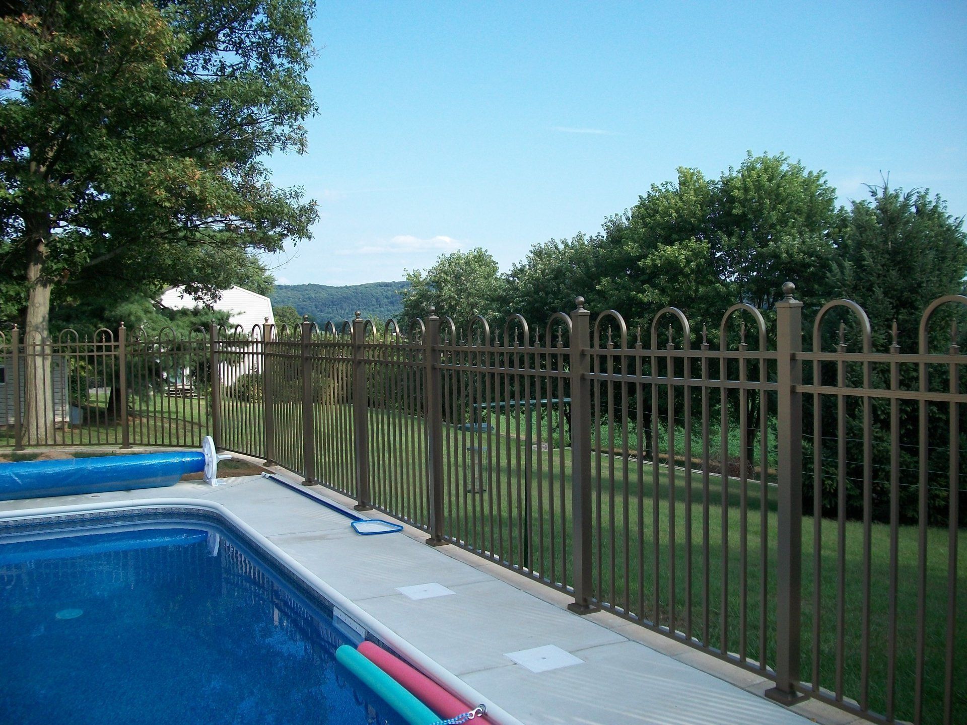Swimming pool with brown metal fence in backyard, trees and blue sky.