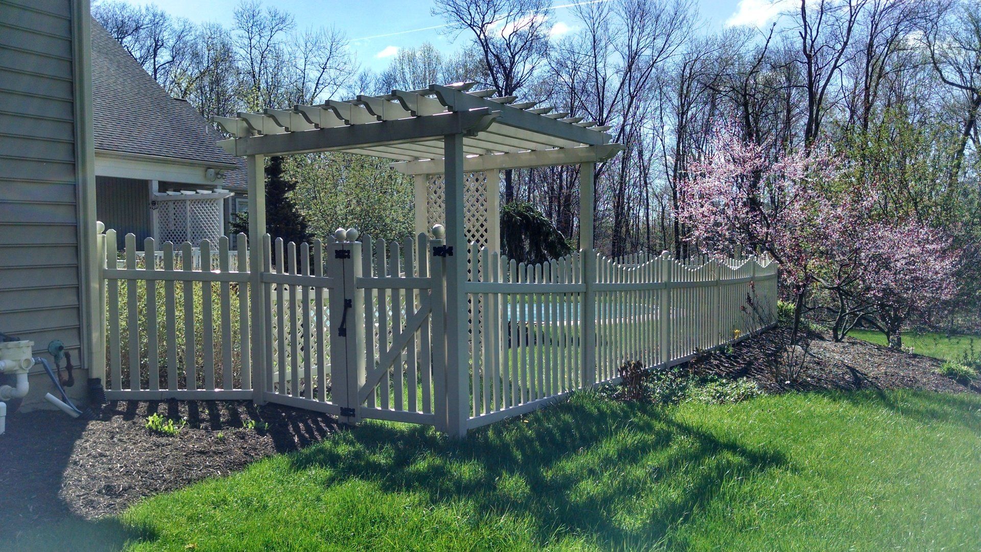 White picket fence with gate, pergola, and flowering tree in a yard.