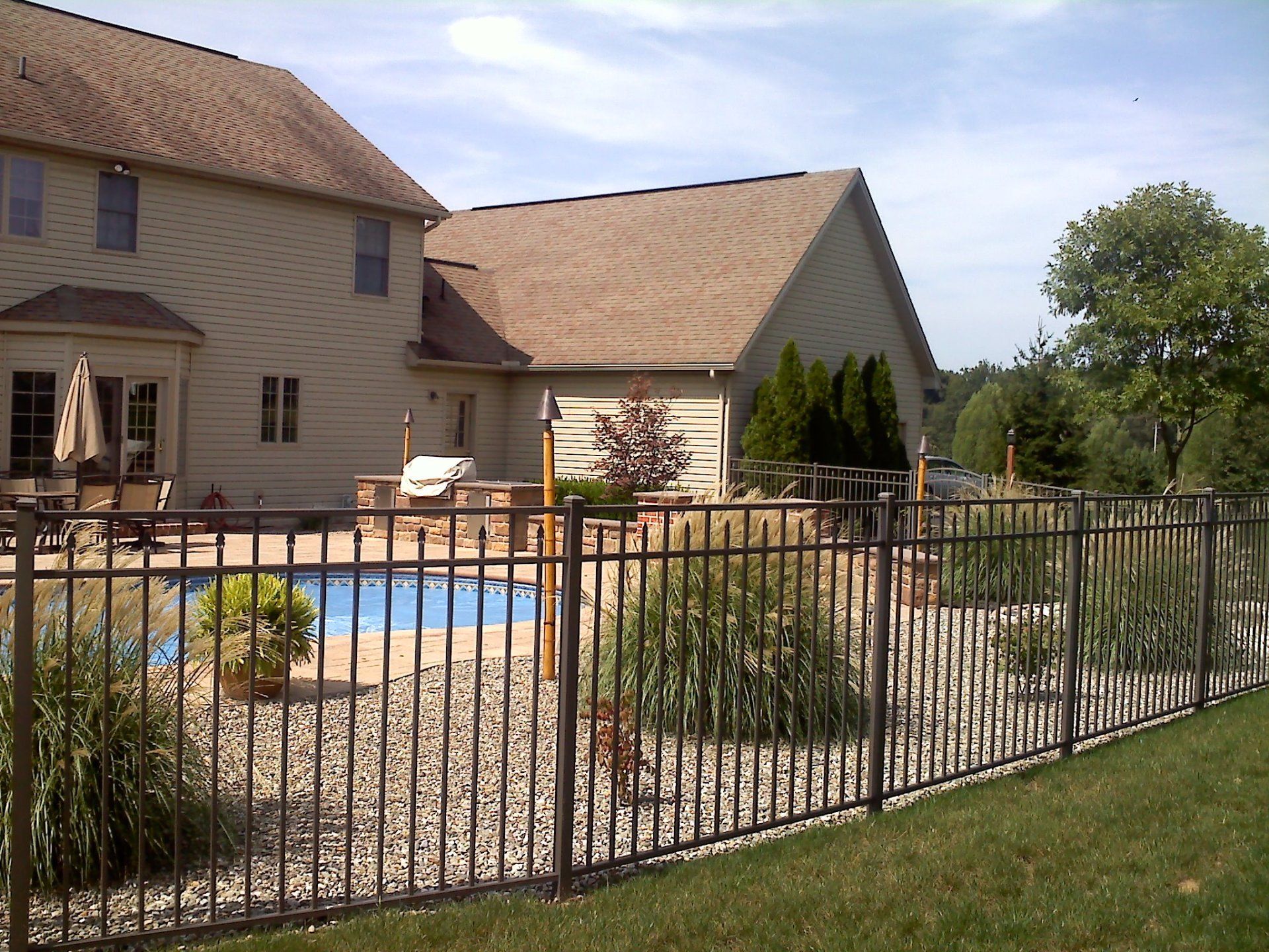 Backyard with a brown house, swimming pool, black fence, and landscaping on a sunny day.