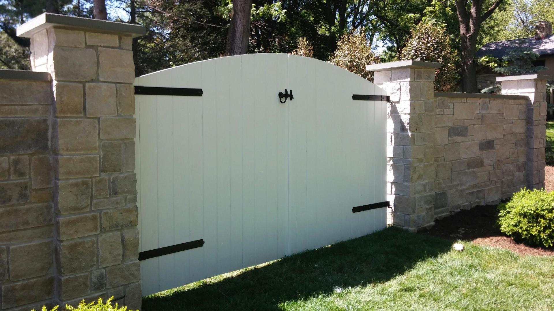 White arched gate with black hardware, flanked by stone walls and pillars, set in a grassy yard.