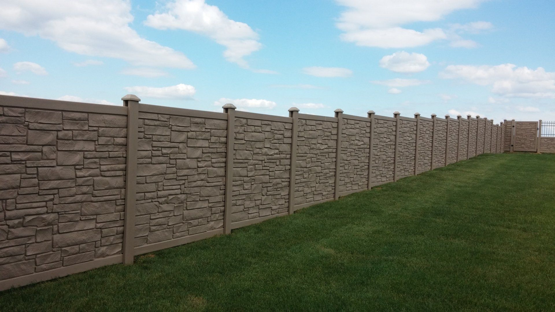 Stone-textured concrete fence in a grassy yard under a partly cloudy blue sky.